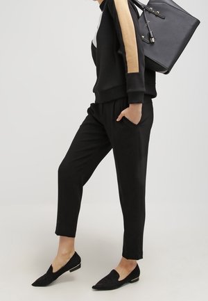 Schwarzer Pullover mit beige farbenen Akzenten, schwarze, zulaufende Hose und schwarze flache Schuhe. Eine schwarze Handtasche wird über der Schulter getragen. Einfaches, lässiges Outfit.