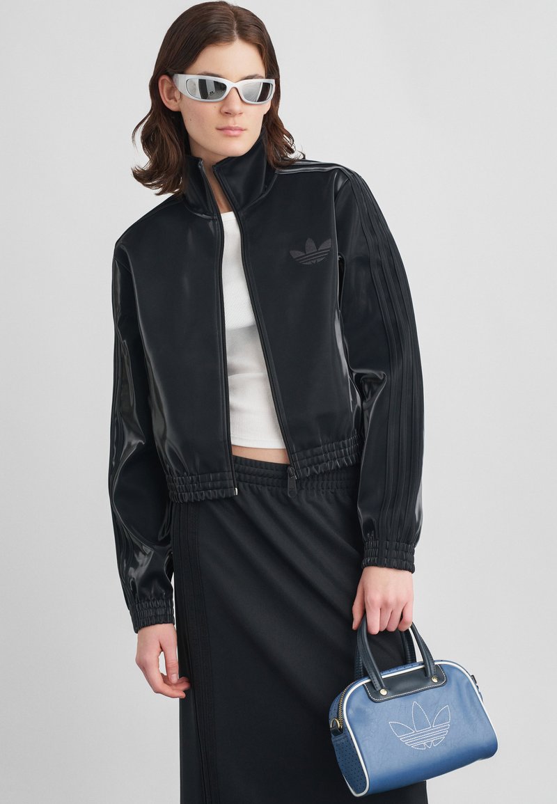 Junge Person, die eine schwarze Reißverschluss-Jacke, einen schwarzen Rock, ein weißes Oberteil und silberne Sonnenbrille trägt, während sie eine blaue Handtasche mit weißem Logo hält.