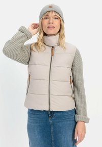 Puffige beige Weste mit strukturierten grauen Strickärmeln, ausgestattet mit einem hohen Kragen, Seitentaschen und braunen Lederakzenten, kombiniert mit einer passenden Mütze.