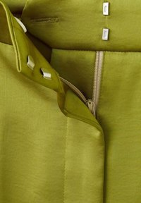 Tejido de satén verde con una textura suave, que presenta herrajes plateados, un cierre de cremallera y detalles de botones en la cinturilla.