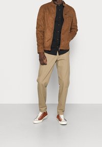 Brun mocka bomberjacka med en zipficka, kombinerad med en svart skjorta med knappar, beige byxor och bruna och vita sneakers.