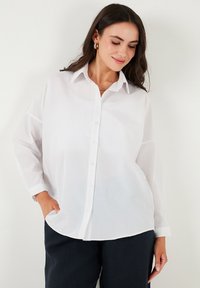 Camicia bianca con bottoni realizzata in tessuto leggero; presenta un colletto, maniche lunghe e un orlo curvo. Abbinata a pantaloni scuri e larghi.