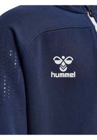 Marineblå atletisk jacka med texturerad yta, vit dragkedja och Hummel-logotyp i vitt. Har ett prickmönster på ena ärmen.