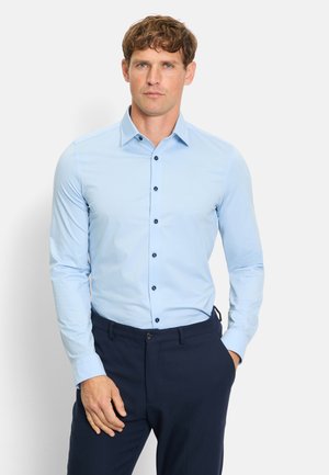 Homme portant une chemise bleu clair à boutons et un pantalon bleu marine foncé, debout avec une main dans la poche devant un fond uni.