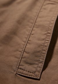 Brun tyg med en strukturerad yta, som har en framträdande sydd söm och en vikt kant. Materialet verkar slätt och följsamt.