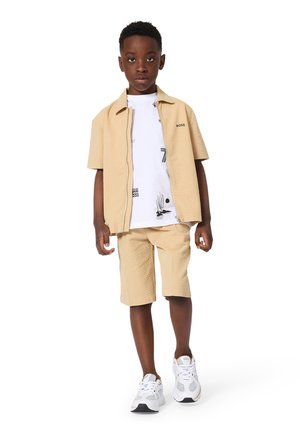 Beige shirtje met korte mouwen en een bijpassende korte broek met een witte grafische T-shirt eronder, gecombineerd met witte sneakers.