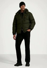 Temno zelena puhovka z kapuco, zadrgo in sprednjimi žepi. Črne hlače in čevlji, kapa na glavi, stoji ob svetli ozadju.