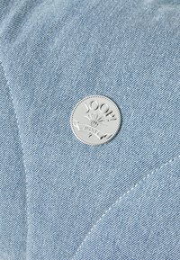 Světle modrá džínovina má šikmé švy a stříbrné kruhové logo s textem "JOOP! JEANS" uprostřed.