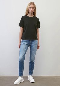 Noor naine musta lühikese varrukaga t-särgiga, helesiniste kulunud teksade ja valgete tossudega, seisab lihtsa helehallseina taustal.