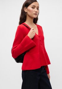 Cardigan tricotat roșu cu decolteu în V, închidere cu nasturi și mâneci largi. Are un tiv evazat și un model texturat. Purtați-l cu blugi închiși la culoare și o geantă neagră.