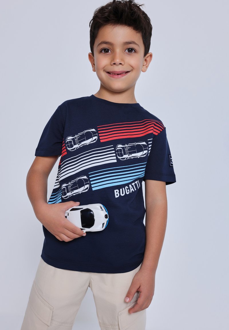 Boy smiling, wearing navy Bugatti t-shirt with red, white, blue racing stripes and car designs, holding white toy car, beige shorts.