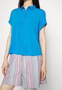 Femme portant une chemise à manches courtes boutonnée bleu vif et un short à rayures verticales multicolores, debout devant un fond blanc uni.