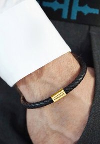 Bracciale in pelle intrecciata nera con chiusura rettangolare dorata incisa con "TISSUVILLE". Indossato su un polso vicino a un polsino di camicia bianco.