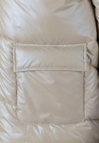 Beige pufferjack stof met een glanzende, gladde textuur; heeft een rechthoekige, gestikte zak aan de linkerzijde.