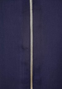 Tela azul marino con una cremallera central de dientes metálicos y una lengüeta de nylon para tirar. Textura suave y diseño limpio.