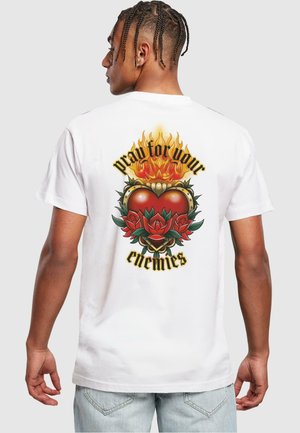 Camiseta blanca de manga corta con un diseño gráfico colorido en la parte posterior, que incluye un corazón, rosas, llamas y el texto "reza por tus enemigos."