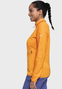 Orange Zip-Jacke mit hohem Kragen, gerippter Textur und einer Seitentasche. Das Model trägt blaue Hosen und schaut nach links.