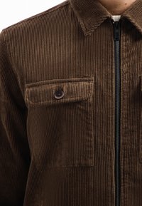 Bruine corduroy jas met een ritssluiting aan de voorkant, een spitse kraag en twee borstzakken met knoopsluitingen; textuur met verticale strepen zichtbaar.
