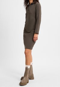Długa sukienka z długim rękawem w oliwkowym kolorze, z kołnierzem na zamek błyskawiczny do połowy, dopasowana do sylwetki, zestawiona z jasnobrązowymi botkami za kostkę i białymi skarpetkami. Gładka tekstura materiału.