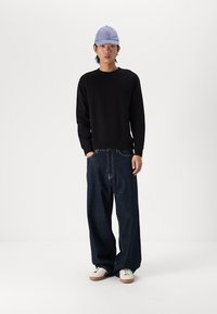 Sudadera negra de manga larga, combinada con pantalones vaqueros de pierna ancha azul oscuro y una gorra azul clara, sobre un fondo blanco liso.