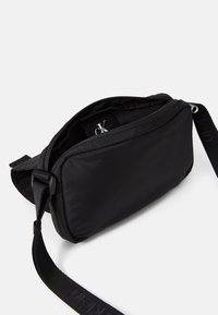 Svart nylon crossover-veske med glidelås, som har en kompakt rektangulær form og en justerbar stropp for enkel bæring.