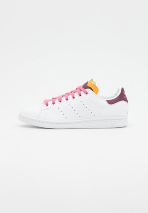 Biały sneaker Adidas z różowymi sznurowadłami, fioletową zakładką na pięcie i pomarańczowym akcentem na języku, prezentowany na jednolitym białym tle.