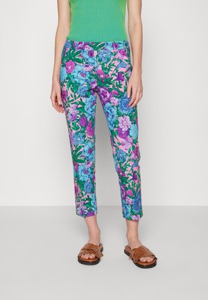 Frau trägt lebhafte Blumenhose in Pink, Blau und Lila, kombiniert mit grünem ärmellosem Top und braunen Sandalen.