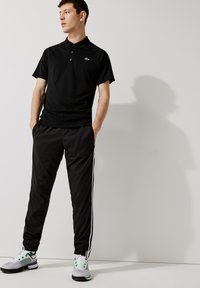 Černé polo tričko s zeleným logem, spárované s černými sportovními kalhotami se bílými bočními pruhy a bílými teniskami se zelenými akcenty.