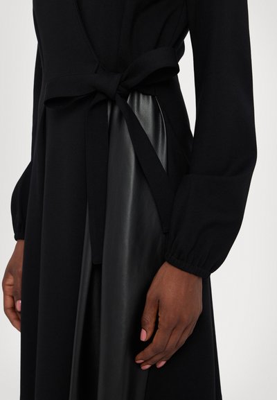 Schwarzes Kleid mit taillierter Oberpartie und lässigen Ärmeln. Verfügt über einen gebundenen Gürtel und ein kontrastierendes glänzendes schwarzes Lederpanel. Weiche Stofftextur.