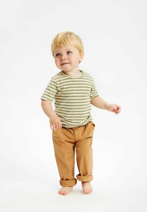 Ljusgrön och vitrandig kortärmad skjorta kombinerad med uppvikta beige byxor. Barnet står barfota, ler och står framför en neutral bakgrund.