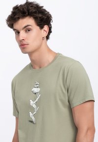 Olijfgroene katoenen t-shirt met een zwart-wit grafisch ontwerp van abstracte figuren die stenen in verticale uitlijning opstapelen.