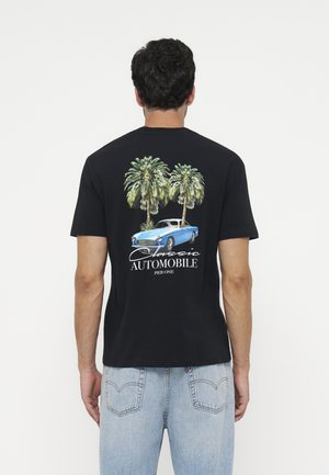 Mand iført sort t-shirt med blå klassisk bil og to palmer grafik og teksten "Classic Automobile Pier One" på ryggen.