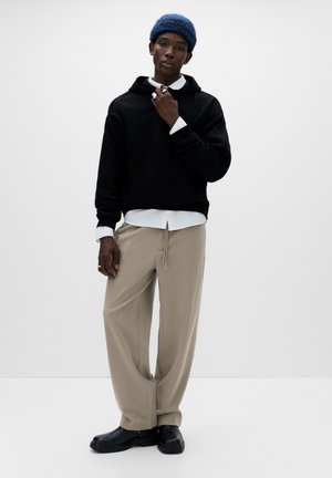 Black hoodie with a kangaroo pocket, beige wide-leg trousers, blue knit beanie, and black shoes, set against a plain white background.