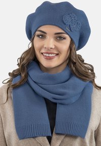 Beret tricotat albastru cu detaliu floral și fular asortat. Fularul are o margine ribbată, ambele fiind realizate dintr-un material moale și texturat.
