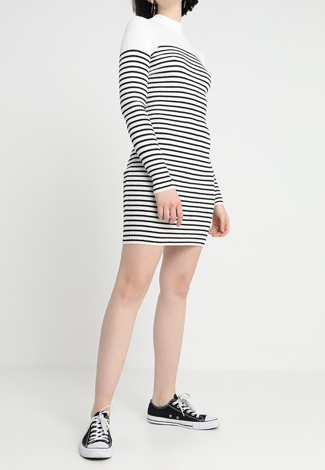 Femme portant une mini-robe à manches longues rayée noir et blanc et des baskets basses noires, debout devant un fond blanc.