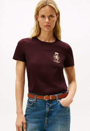 T-shirt di cotone color bordeaux con collo a girocollo, caratterizzata da un emblema dorato sul petto sinistro. Abbinata a jeans in denim blu e una cintura marrone.