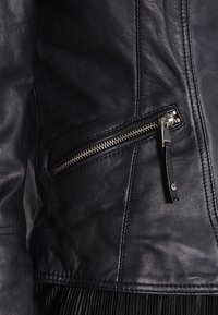 Černá kožená bunda s diagonální zipovou kapsou, stříbrnými detaily a texturovanou tkaninou se švy. Pás s třásněmi.