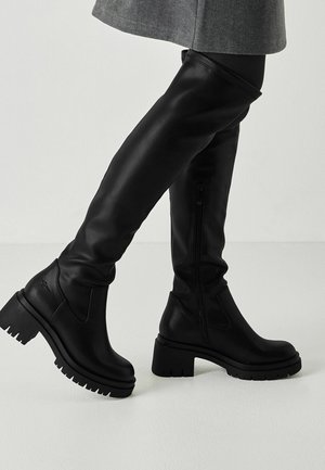 Bottes en cuir noires montantes jusqu'aux genoux avec talons épaissis et fermetures éclair latérales portées avec un pantalon gris. Jambes croisées sur un fond clair.
