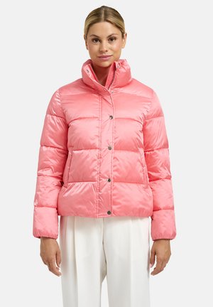 Frau trägt eine glänzende pinke Steppjacke mit hohem Kragen und Druckknöpfen, kombiniert mit weißen weiten Hosen, stehend vor weißem Hintergrund.