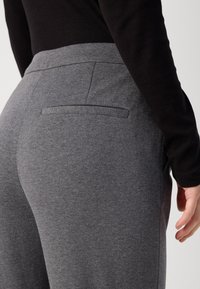Un pantalon gris en maille présente une texture lisse avec une ceinture élastique et une unique poche arrière, montré de profil avec un haut noir.