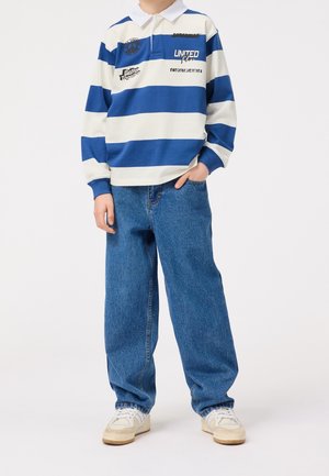 Blau-weiß gestreiftes Polohemd mit langen Ärmeln und Kragen; kombiniert mit lockeren blauen Jeans und weißen Turnschuhen. Verfügt über gestickte Logos.