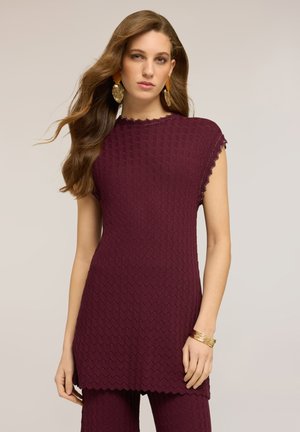 Kvinde med langt brunt hår iført en ærmeløs, tekstureret, mahognifarvet top og matchende bukser, accessoriseret med store guldsmykker og armbånd.