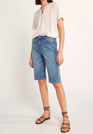 Kvinde iført en hvid kortærmet bluse, blå knælange jeansshorts og brune sandaler med stropper og flad sål, stående mod en ensfarvet baggrund.