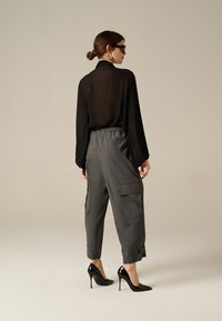 Blusa nera trasparente a maniche lunghe abbinata a pantaloni cargo grigi con fascia elastica in vita e tasche, completata da scarpe nere con tacco alto.