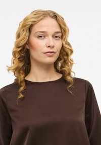 Brūns satīna blūze ar garām piedurknēm, apaļu izgriezumu un gludu tekstūru. Raksturo smalka spīduma un minimālistisks dizains.