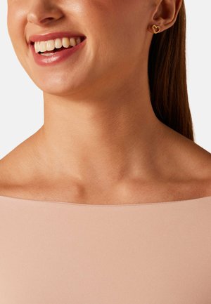 Close-up van een glimlachende vrouw die gouden oorstekers in de vorm van een hart draagt en een nude off-shoulder top tegen een effen achtergrond.