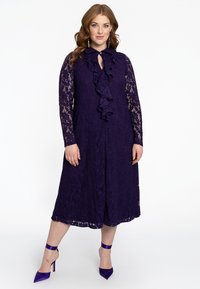 Paarse kanten jurk met lange mouwen, gerimpelde voorkant, lengte tot de knie. Gecombineerd met paarse hoge hakken met enkelbandjes.