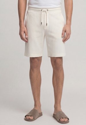 Uomo che indossa pantaloncini a coste color avorio con coulisse e sandali taupe slip-on, in piedi davanti a uno sfondo chiaro e semplice.