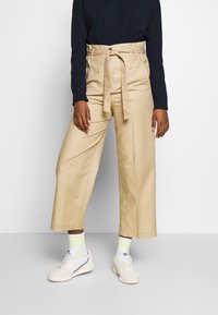 Beige weite Hosen mit hoher Taille und Gürtel, aus glattem Stoff. Kombiniert mit weißen Sneakers und gestreiften Neon-Socken.