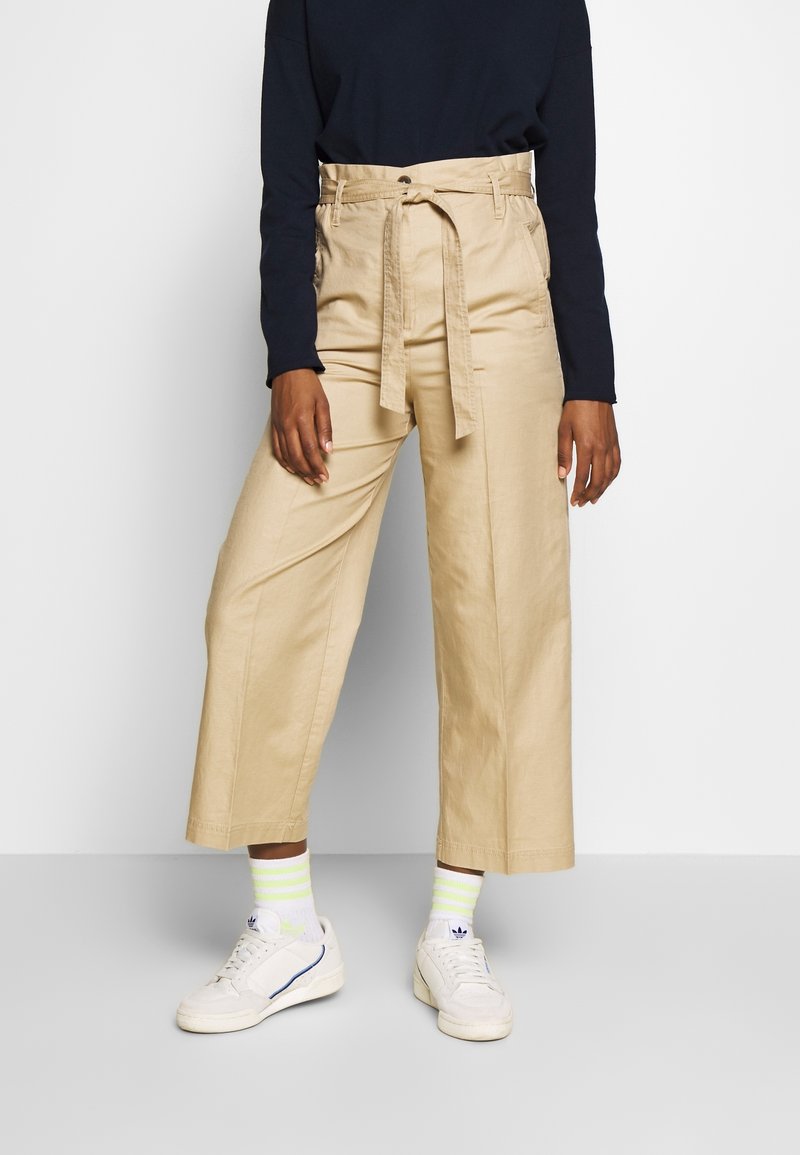 Beige weite Hosen mit hoher Taille und Gürtel, aus glattem Stoff. Kombiniert mit weißen Sneakers und gestreiften Neon-Socken.